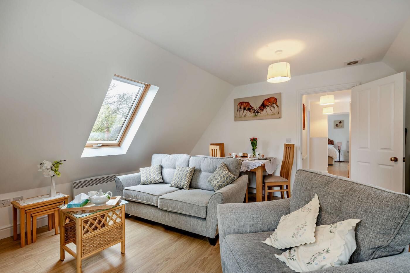 Un salon chaleureux avec un canapé gris, une table à manger avec des chaises, une petite table d’appoint et une fenêtre donnant sur des arbres.