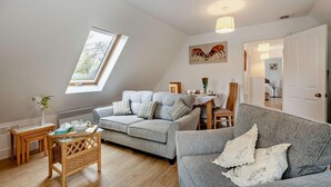 A cozy living room with a grey sofa, a dining table with chairs, a small side table, and a window with a view of trees. - 2 Bed in Eccles (oc-l27507) (Eccles)
