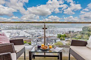 A balcony with wicker furniture, a table with breakfast items, and a view of a marina and town. - 3 Bed in Kingswear (oc-bc121) (Dartmouth)