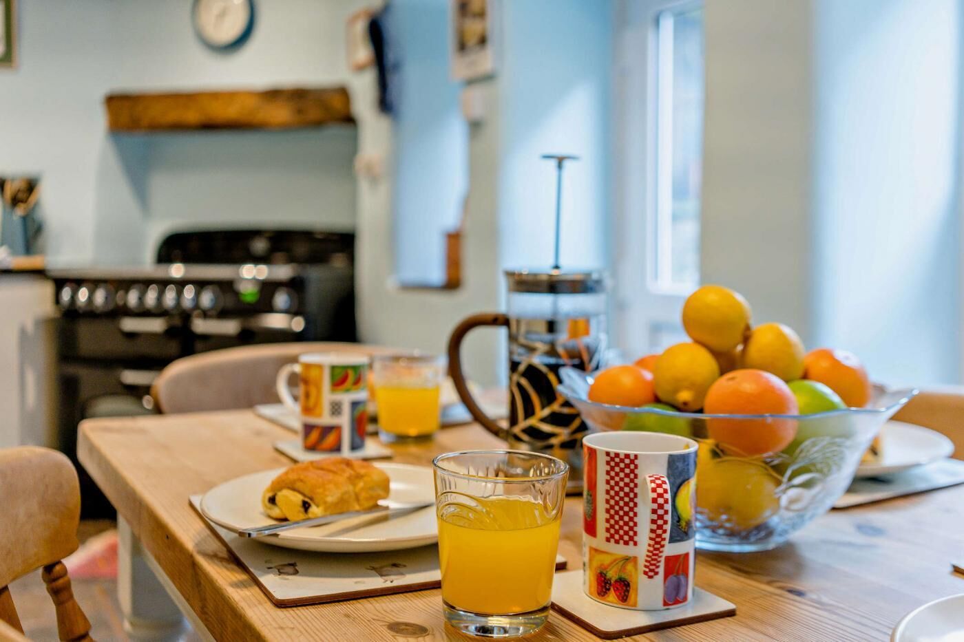 Uma mesa posta para o café da manhã, com uma tigela de frutas, uma xícara de café e um copo de suco de laranja.