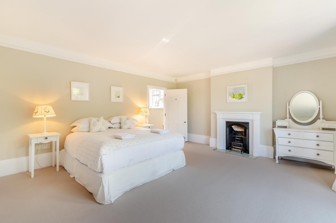 A spacious bedroom with a large bed, bedside tables, a dresser, a mirror, and a fireplace.