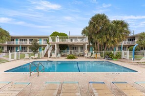 A pool area with lounge chairs, a lifeguard stand, and a two-story building with balconies. - Pompano 3225 (Pompano Beach)