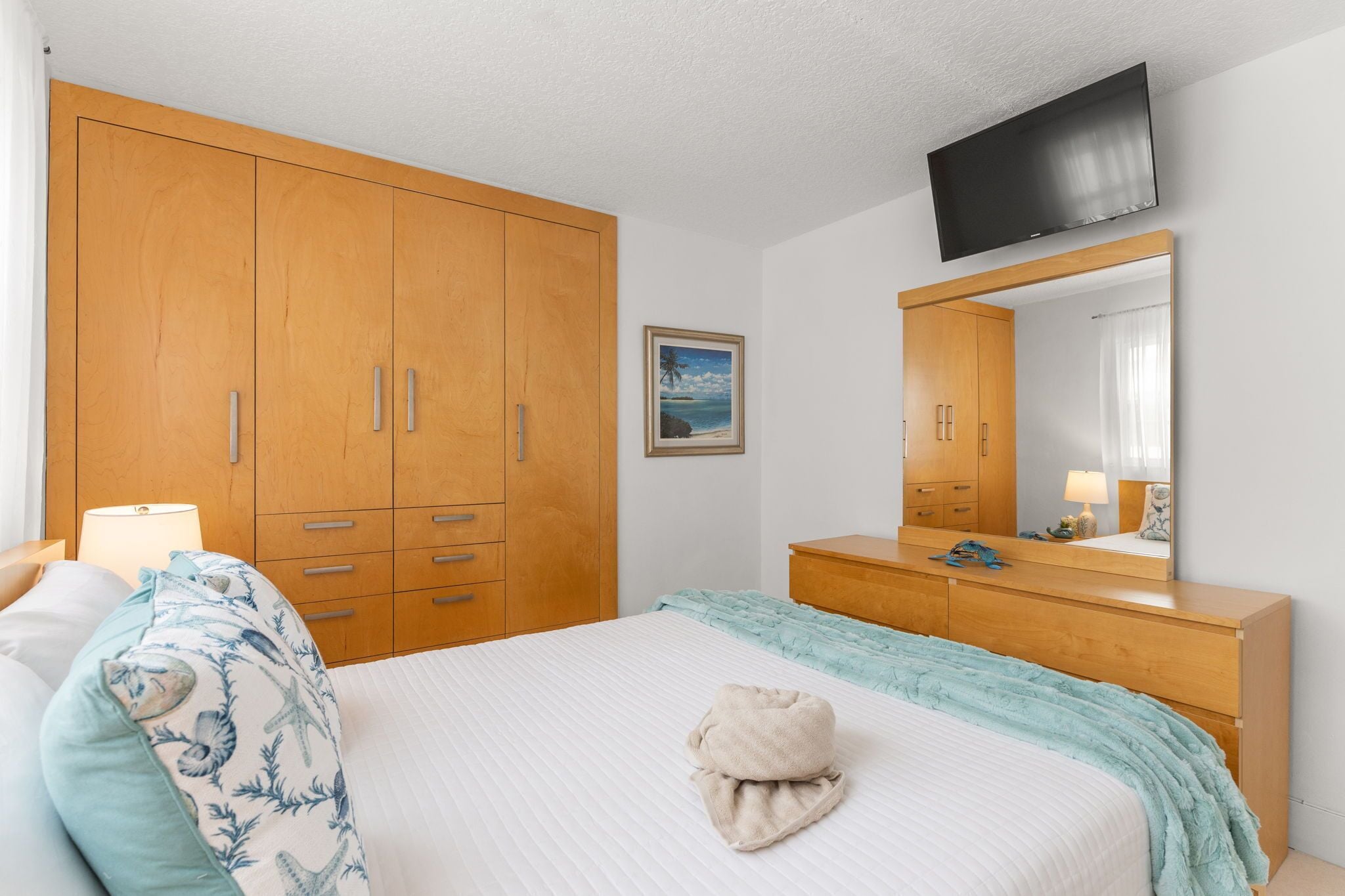 Une chambre avec un grand lit, une armoire en bois, un téléviseur fixé au mur et un tableau encadré accroché au mur.