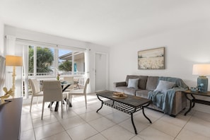 A living room with a sofa, coffee table, and chairs. There is a balcony with a view of palm trees. - Pompano 3225 (Pompano Beach)