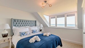 A bedroom with a tufted headboard, a large window with a view of the sea, and a ceiling light fixture. - 3 Bed in Seahouses (oc-p27534) (Seahouses)