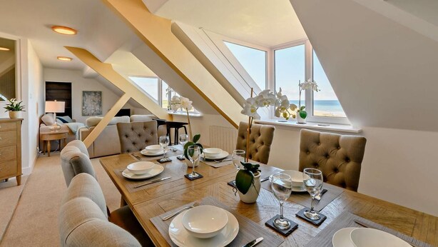 A dining area with a wooden table set for four, a sofa, and a view of the beach through large windows. - 3 Bed in Seahouses (oc-p27534) (Seahouses)