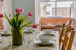 A dining table set with white plates, bowls, and wine glasses, a vase with pink tulips, and a view of a wooden deck and a car through a glass door.
