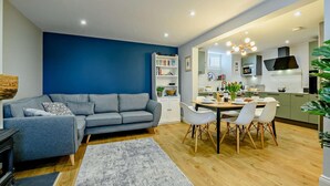A modern living room with a grey sofa, a dining table with chairs, and a kitchen area with a stove and cabinets. - 3 Bed in Ruswarp (oc-wa264) (Ruswarp)