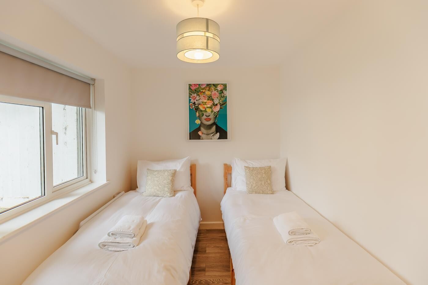 Ein Zimmer mit zwei Einzelbetten, einer Deckenleuchte, einem Fenster und einem an der Wand hängenden Bild.