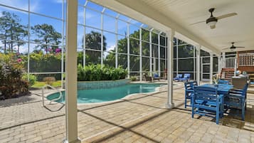 A covered pool area with a clear view of the pool, surrounded by blue chairs and a table, and a ceiling fan.