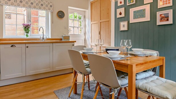 Una cocina con armarios blancos, una mesa de comedor de madera dispuesta para cuatro personas y una alfombra estampada.