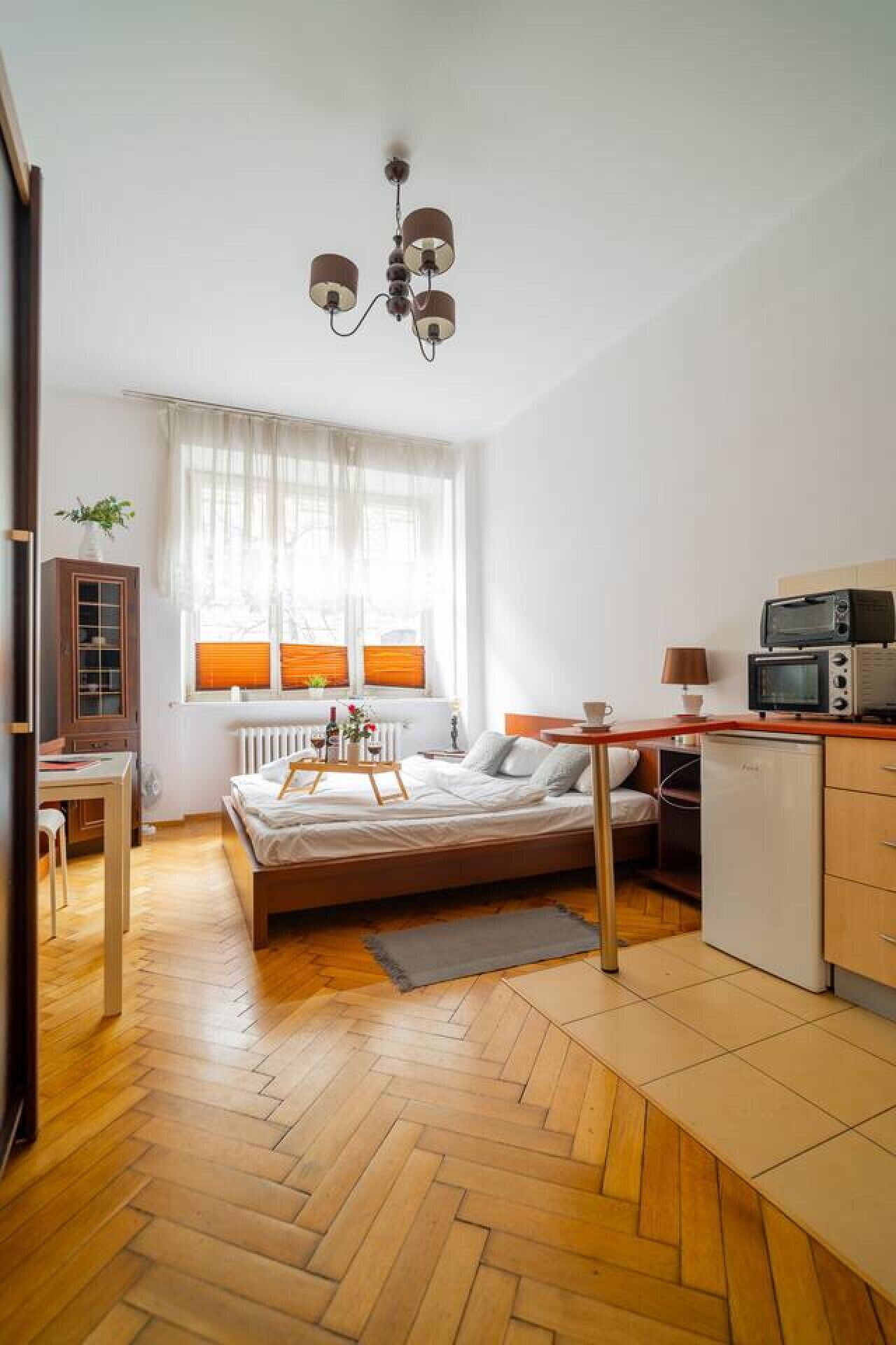 Una stanza compatta con un letto, un angolo cottura, pavimento in legno e una finestra con tende.