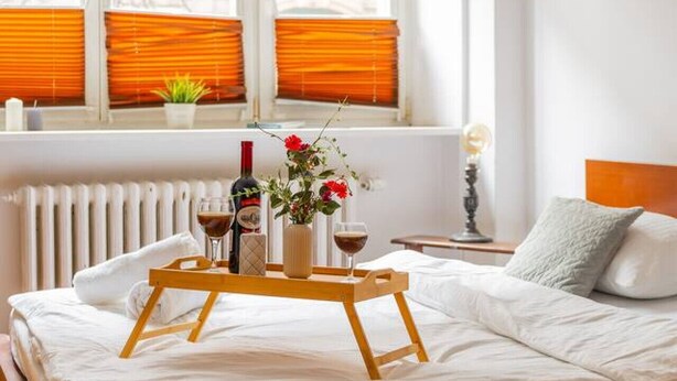 Un letto rifatto con cura, un vassoio con bevande e fiori, un comodino con una lampada e una finestra con persiane arancioni.