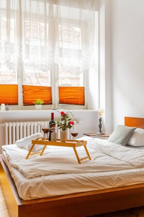 A neatly made bed with a tray of drinks and flowers, a bedside table with a lamp, and a window with orange blinds. - Batorego Studio - Old Town nearby - by Rentujemy (Kraków)