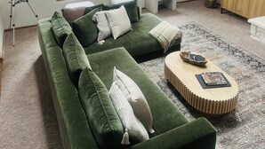 Un sofá verde con varios cojines, una mesa de centro y un telescopio en un salón con vistas a muebles de exterior a través de amplias ventanas.