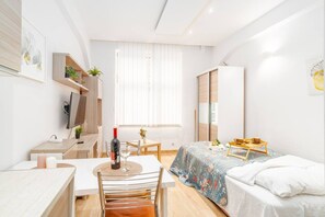Ein modernes, helles Schlafzimmer mit Bett, Schreibtisch und Stuhl. Ein Fenster mit Jalousien, ein Kleiderschrank und ein kleiner Tisch mit Tablett sind ebenfalls vorhanden.