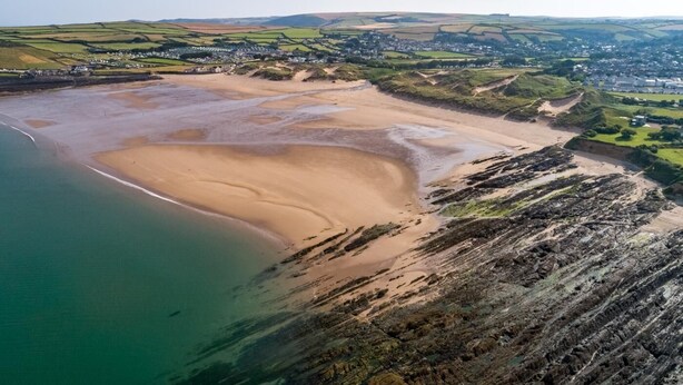 Vue aérienne d’une zone côtière avec une plage de sable, des falaises rocheuses et une petite ville en arrière-plan.