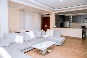A modern living room with a grey sectional sofa, a white coffee table, and a kitchen area with dark cabinets and a microwave. - One-Bedroom Apartment #4 (Kigali)