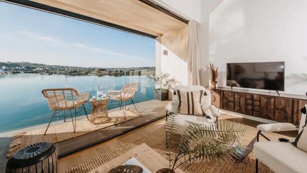 A modern living room with a large window overlooking a body of water, a flat-screen TV, and a dining area with wicker furniture. - 3 Bed in Hayle (oc-t27597) (Hayle)