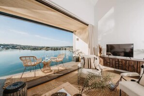 Ein modernes Wohnzimmer mit einem großen Fenster mit Blick auf ein Gewässer, einem Flachbildfernseher und einem Essbereich mit Korbmöbeln.