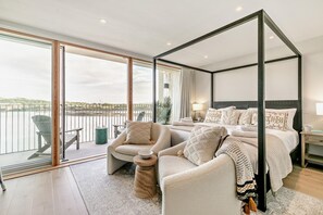 A modern bedroom with a four-poster bed, a seating area with a chair and a small table, and a balcony with a chair and a trash can. - 3 Bed in Hayle (oc-t27597) (Hayle)
