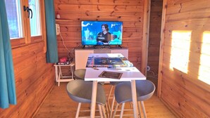 Une pièce de cabane en bois comprenant une télévision, une petite table, deux chaises et une fenêtre avec des rideaux.