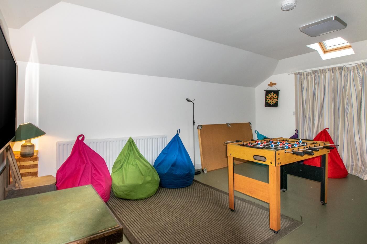 Une pièce chaleureuse avec un baby-foot, des poufs colorés, une lampe et un jeu de fléchettes.