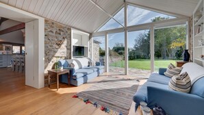 A spacious living area with a stone wall, a blue sofa, a wooden coffee table, and a view of a garden through large windows. - 3 Bed in St. Minver (oc-litmow) (Saint Minver)