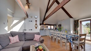 A cozy living room with a grey sofa, a dining area with a table set for four, and a kitchen in the background. - 3 Bed in St. Minver (oc-litmow) (Saint Minver)
