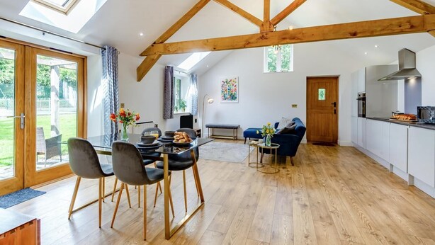 A spacious living area with a high ceiling and exposed wooden beams, a dining area with modern furniture, and a kitchen with white cabinets and a stainless steel range hood. - 1 Bed in Dunston (oc-h27629) (Stoke Holy Cross)