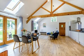 Un vaste espace de vie doté d’un haut plafond et de poutres apparentes en bois, un coin repas avec des meubles modernes, et une cuisine avec des armoires blanches et une hotte de cuisinière en acier inoxydable.