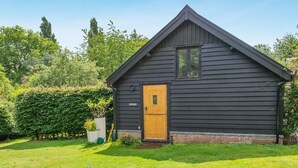 A small, dark wooden cabin with a single door and window, surrounded by a well-maintained lawn and dense greenery. - 1 Bed in Dunston (oc-h27629) (Stoke Holy Cross)