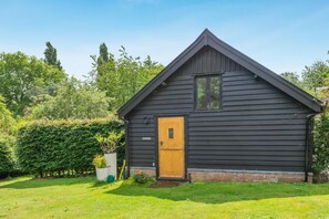Une petite cabane en bois sombre, avec une seule porte et une seule fenêtre, entourée d’une pelouse bien entretenue et d’une végétation luxuriante.