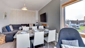 Ett modernt vardagsrum med en matplats, en grå soffa, en tv som är monterad på väggen och ett stort fönster med utsikt över hus.