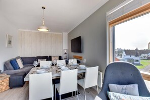 A modern living room with a dining area, a grey sofa, a TV mounted on the wall, and a large window offering a view of houses.