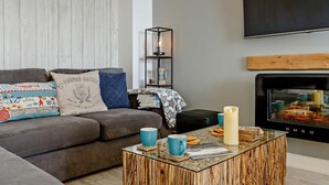 Ett vardagsrum med en grå soffa, mönstrade kuddar, ett soffbord i glas, en blå mugg och en öppen spis.