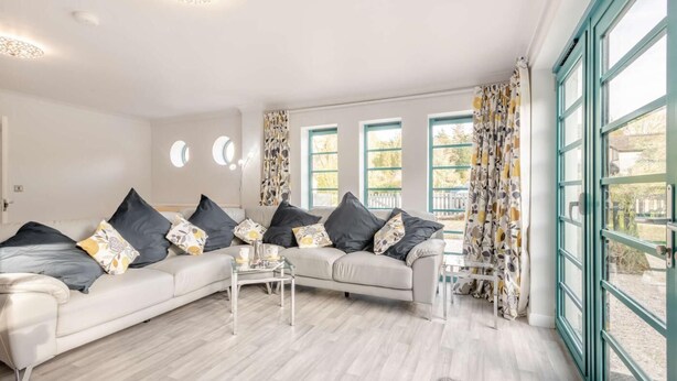A modern living room with a large L-shaped sofa, a coffee table, and a glass-top table. There are three windows with curtains, and a view of the outside. - 2 Bed in St. Olaves (oc-t27605) (Haddiscoe)