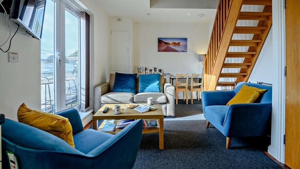 Ein modernes Wohnzimmer mit einer Couch, Sesseln, einem Couchtisch und einem Essbereich mit Tisch und Stühlen. Im Hintergrund ist eine Holztreppe zu sehen.