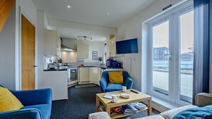 Ein modernes Wohnzimmer mit Sofa, Couchtisch und Fernseher. Es gibt eine Küchenzeile mit Herd und Schränken.