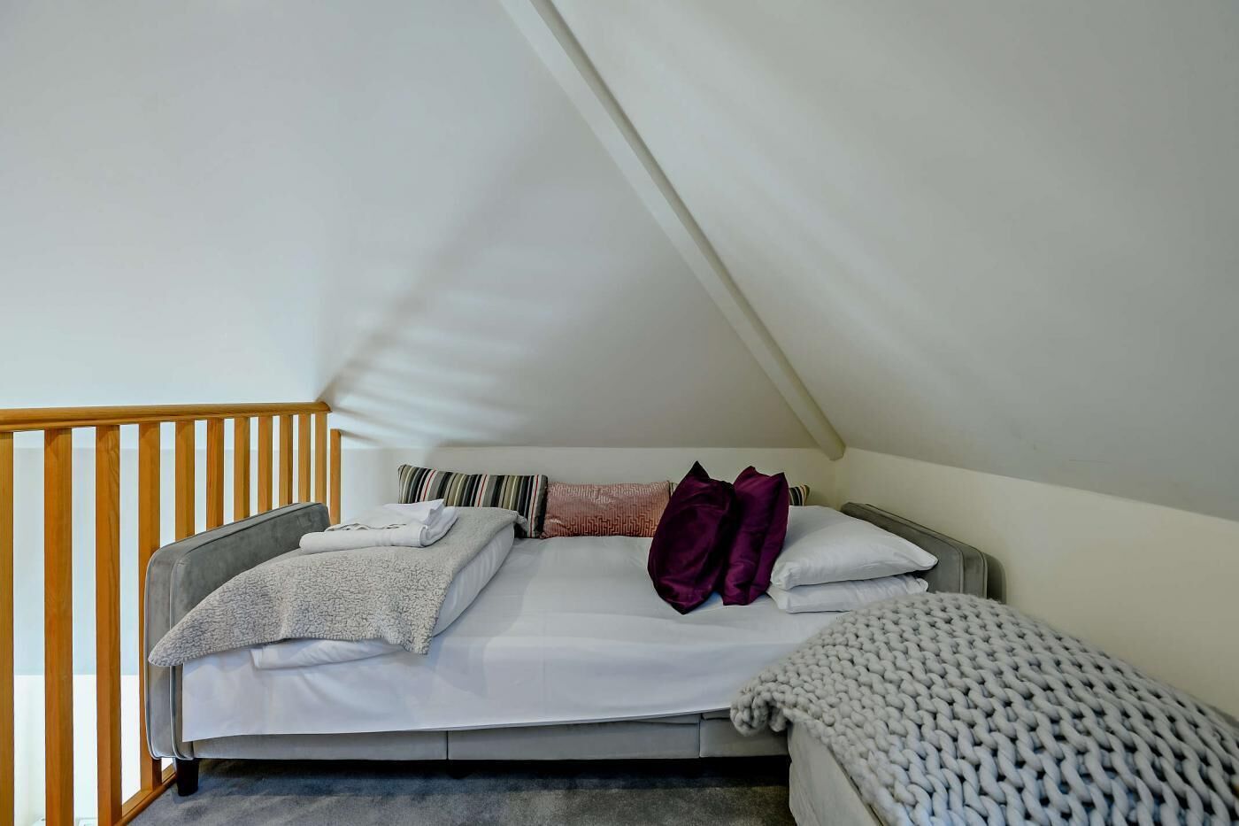Een slaapkamer met een schuine kap, een bed met meerdere kussens en een dekbed, een houten leuning en een klein tafeltje.
