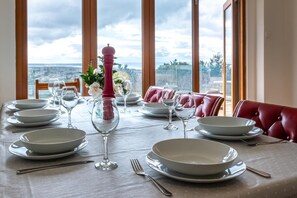 Ein Esstisch für vier Personen, gedeckt mit weißen Tellern, Weingläsern und einem rosa Salzstreuer. Im Hintergrund ist eine schöne Aussicht durch große Fenster zu sehen.