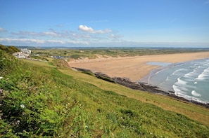 Eine Küstenlandschaft mit Sandstrand, grasbewachsenen Klippen und einem Gebäude in der Ferne.