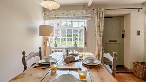 A dining room with a wooden table set for breakfast, a window with floral curtains, and a view of greenery outside. - 2 Bed in Southrepps (oc-n27613) (Southrepps)