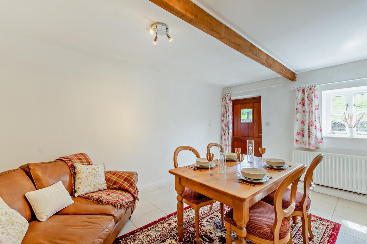 A cozy living room with a brown leather sofa, a wooden dining table set for four, and a red patterned rug.