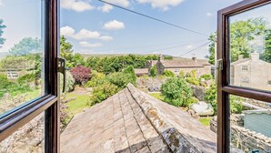 A view from a window overlooking a stone roof, a garden with various plants, and traditional stone buildings in the background.