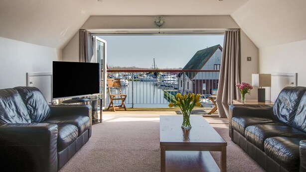 Een ruime woonkamer met een grote televisie, leren banken, een salontafel en een balkon met uitzicht op boten en gebouwen.