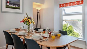 Un comedor con una mesa de madera dispuesta para cuatro personas, rodeada de sillas negras. Un jarrón con girasoles está sobre la mesa, y hay una ventana con vistas a tejados.