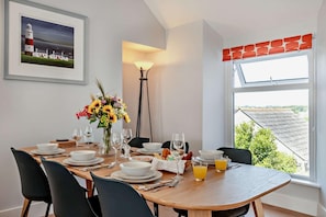 A dining area with a wooden table set for four, surrounded by black chairs. A vase with sunflowers is on the table, and there is a window with a view of rooftops. - 3 Bed in Tenby (oc-c27685) (Tenby)