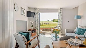 Una sala de estar moderna con un sofá, una mesa de comedor preparada para dos personas, un televisor montado en la pared y un amplio ventanal con vistas a un campo y una valla.