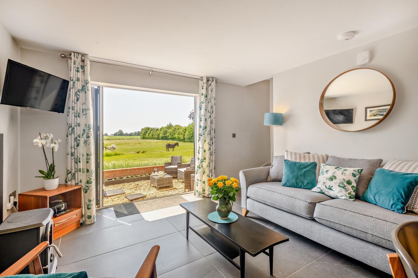 A modern living room with a grey sofa, a flat-screen TV, a coffee table, and a view of a green field with cows.